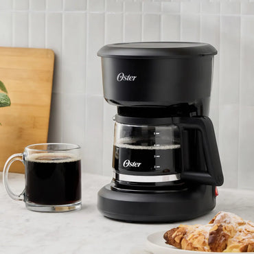 cafetera eléctrica Oster 5 tazas negra con jarra de vidrio taza de café y pastel sobre encimera