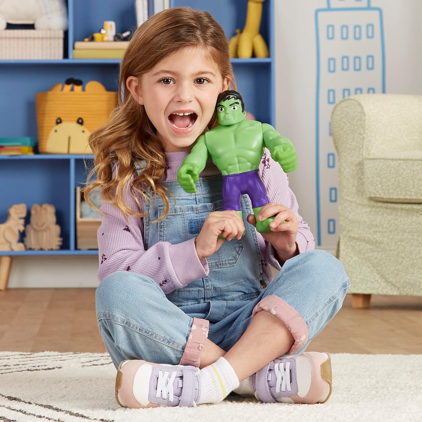 Muñeco Hulk verde con ropa morada, niña sentada en el suelo, habitación colorida.