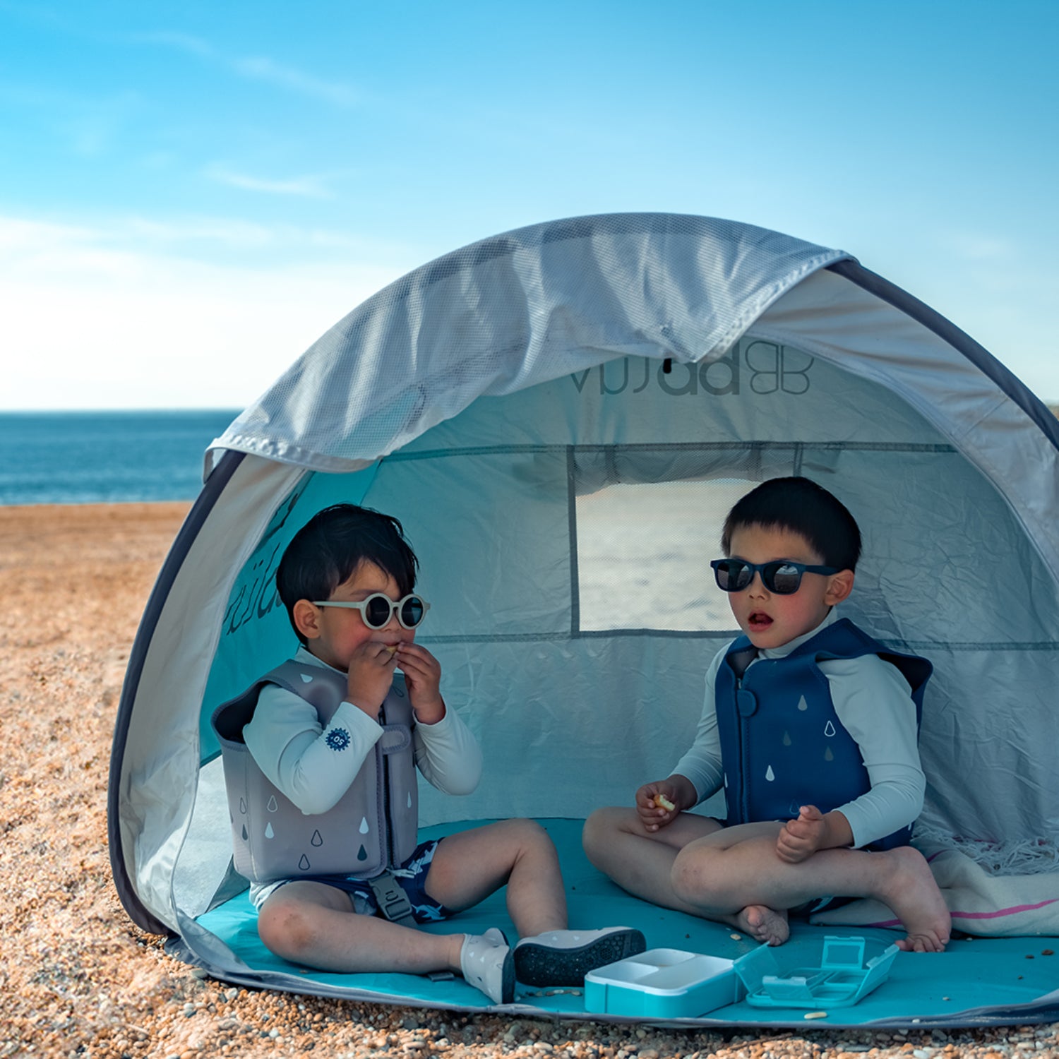 Chaleco BBLUV azul para niños, en carpa en la playa, disfrutando del sol.