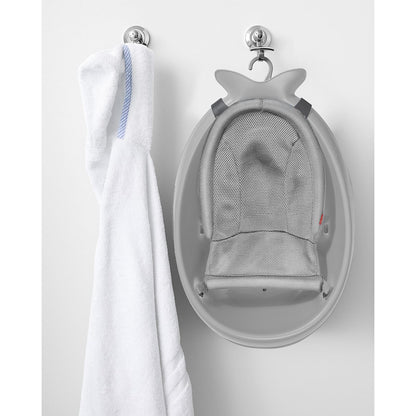 Bañera con hamaca para bebé gris colgada en la pared, con toalla blanca al lado