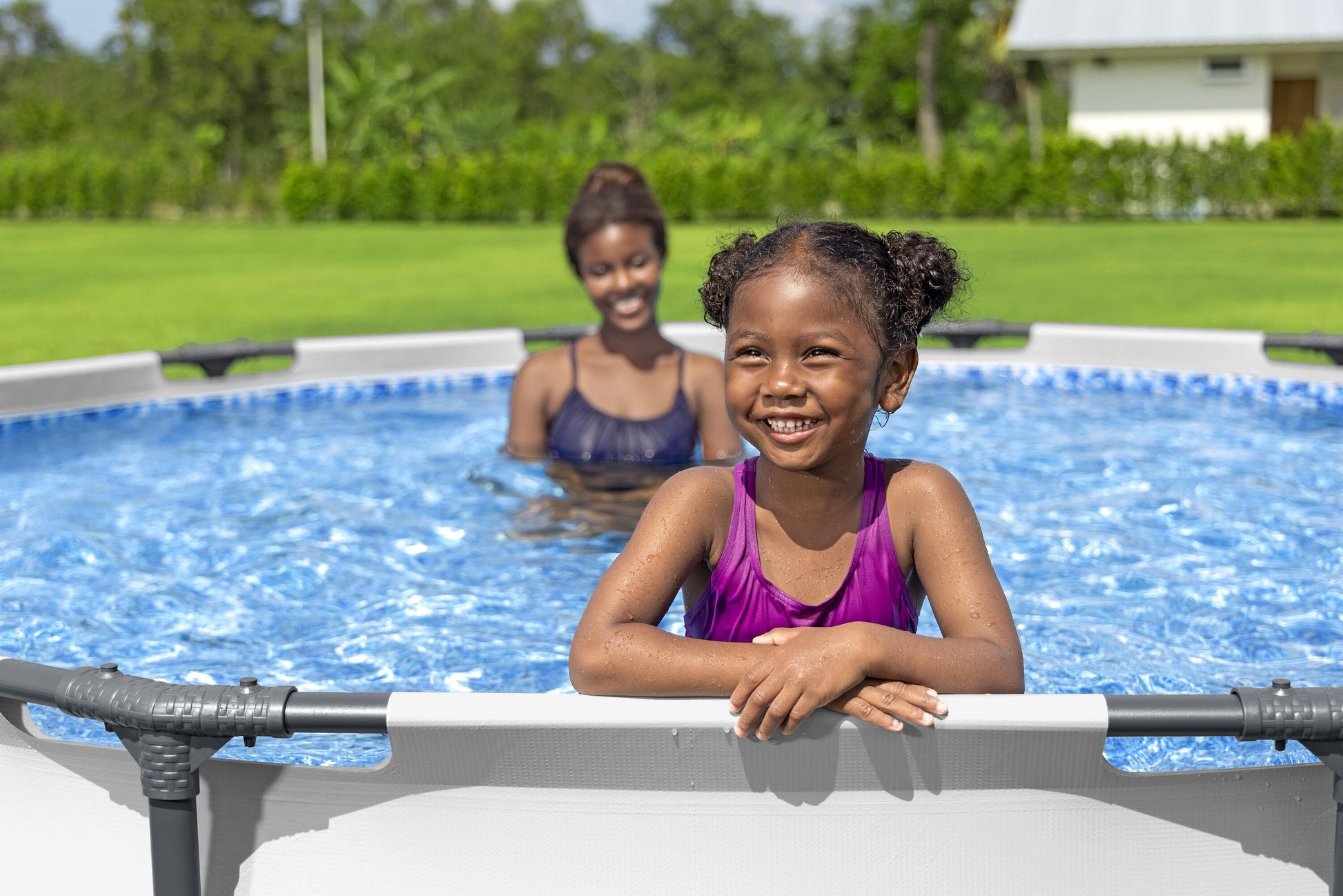 Niña sonriente en piscina desmontable redonda 3.66x0.76 metros, mamá en el fondo, jardín verde.