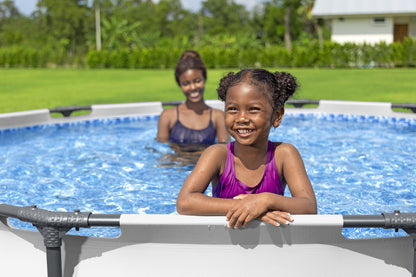 Niña sonriente en piscina desmontable redonda 3.66x0.76 metros, mamá en el fondo, jardín verde.