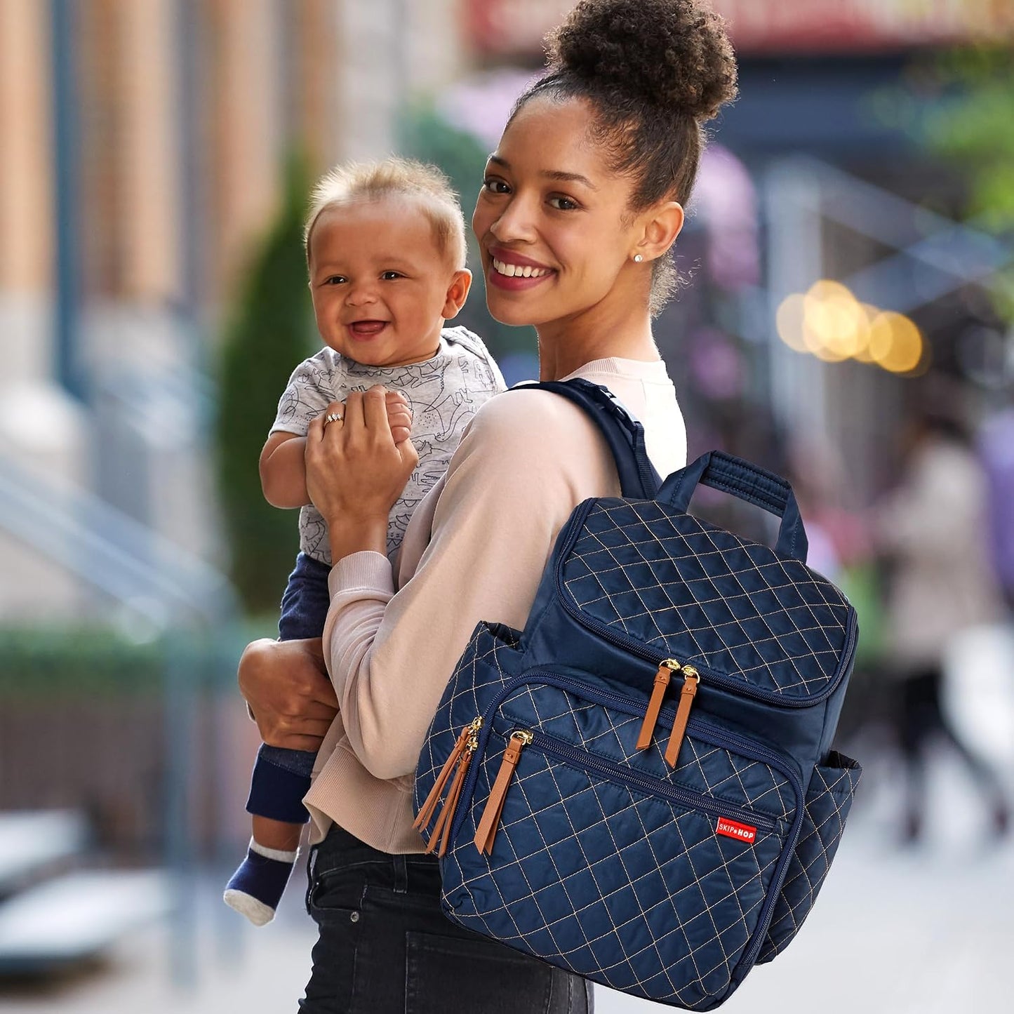 Mochila SKIP*HOP azul con compartimentos, sostenida por madre con bebé