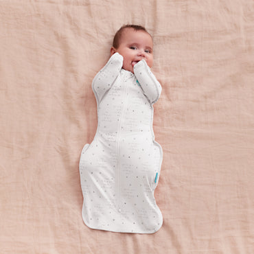 Swaddle blanco de LOVE TO DREAM, vista superior, bebé sonriendo sobre sábanas.